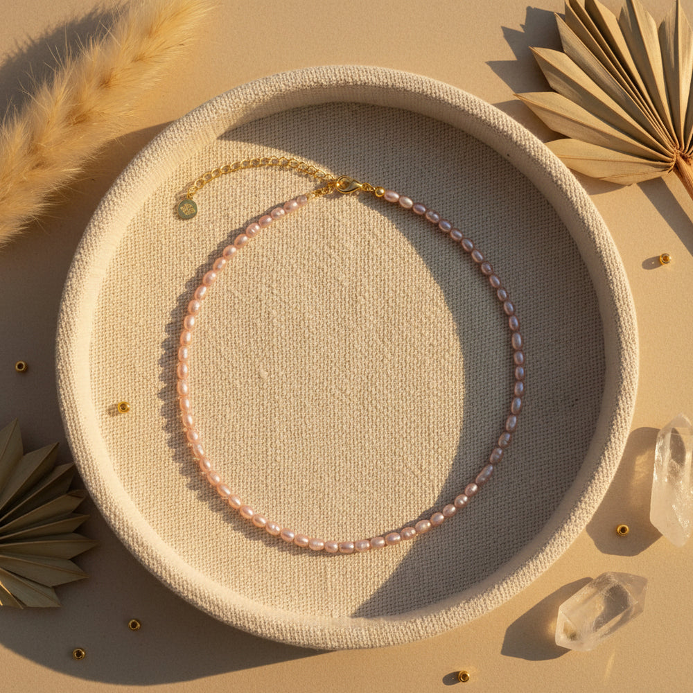 A delicate necklace made of small, light, irregularly shaped pink freshwater pearls arranged in a circle, finished with a gold-tone lobster clasp and an adjustable gold-tone chain extender with a small round charm, shown on a solid white background.